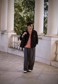 Άτομο με μαύρο μπουφάν πάνω από ροζ πουκάμισο, γκρίζες φαρδιές παντελόνες και λευκά παπούτσια· κρατάει μια μαύρη τσάντα, φοράει γυαλιά ηλίου και στέκεται δίπλα σε κολώνες.