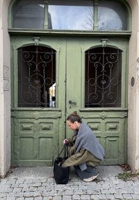 Bolso tote de cuero negro sobre adoquines, junto a puertas de madera verdes con rejas metálicas decorativas y texturas desgastadas. Tela gris colgada sobre los hombros.