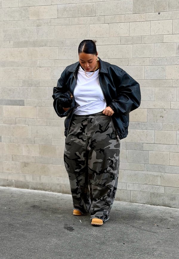 Kvinne iført svart skinnjakke, hvit t-skjorte, kamuflasjebukser og beige støvler, stående mot en lys flislagt vegg, og ser ned.