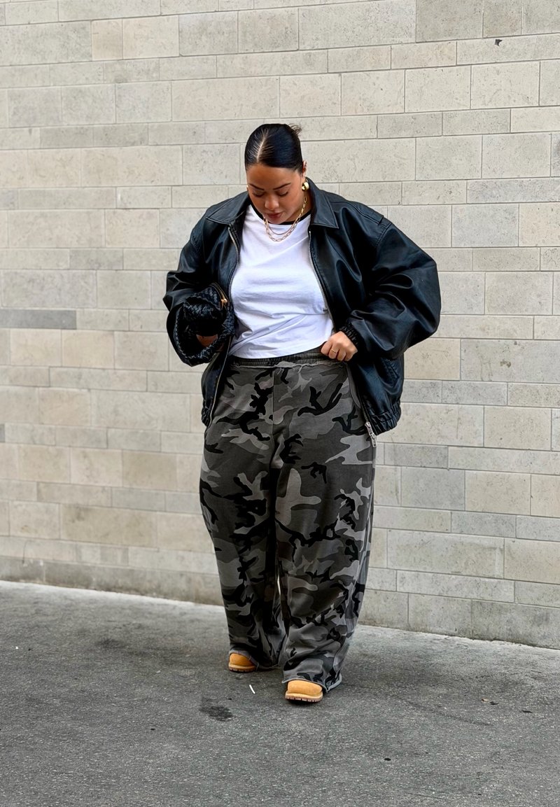 Woman wearing a black leather jacket, white t-shirt, camo pants, and tan boots, standing against a light tiled wall, looking down.