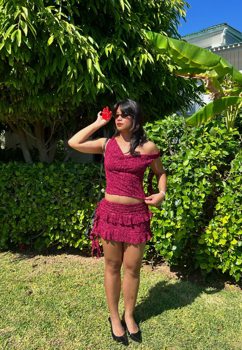 Bordeaux cropped top en gerimpelde minirok van gestructureerde stof, gecombineerd met zwarte hakken. Vergezeld van een rode bloemenaccessoire.