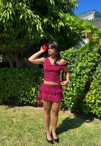 Bordeaux cropped top en gerimpelde minirok van gestructureerde stof, gecombineerd met zwarte hakken. Vergezeld van een rode bloemenaccessoire.