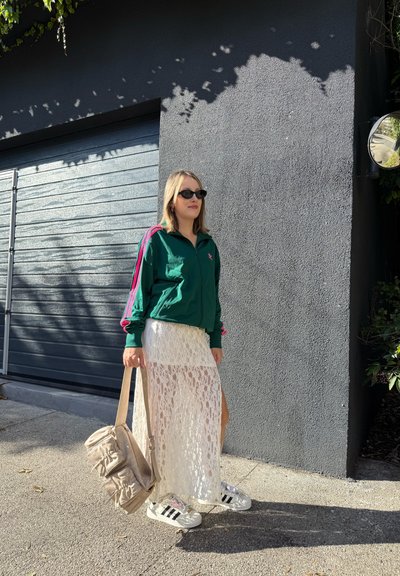 Mujer con gafas de sol, chaqueta deportiva verde, falda de encaje blanca y zapatillas sosteniendo un bolso beige, de pie junto a una pared gris y una puerta de garage.