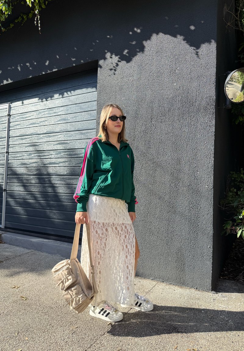 Femme portant des lunettes de soleil, une veste de sport verte, une jupe en dentelle blanche et des baskets, tenant un sac beige, se tenant près d'un mur gris et d'une porte de garage.