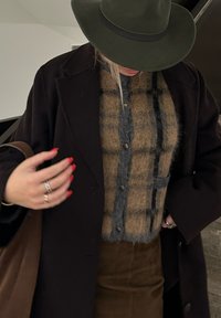 Καφέ πλεκτό πουλόβερ με καρώ σχέδιο και γκρι λεπτομέρειες, μαύρο παλτό, πράσινη καπέλο με φαρδύ σώμα και καφέ φούστα. Χέρια με δαχτυλίδια και κόκκινο μανικιούρ.