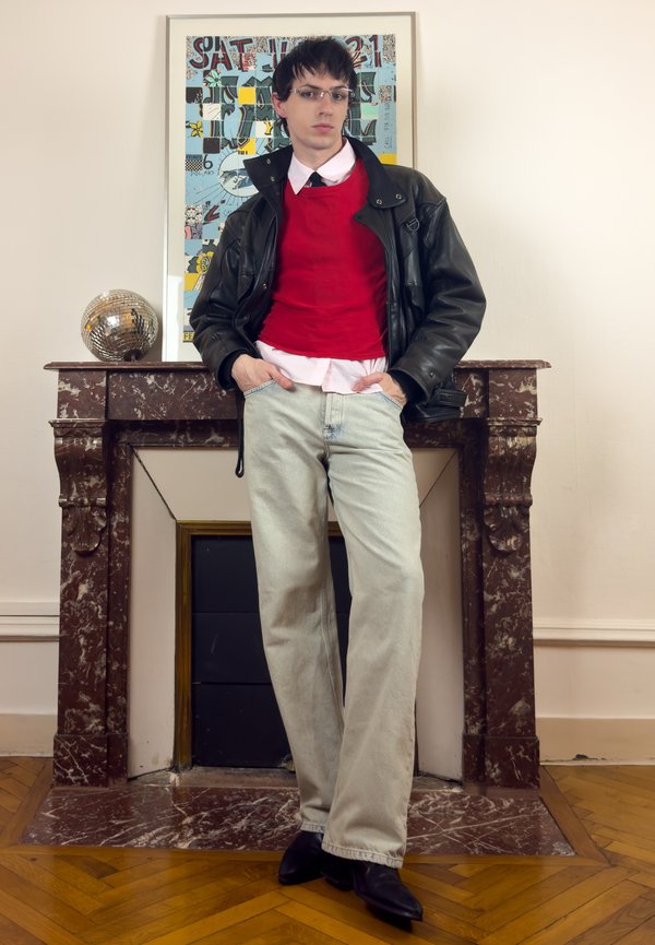 Giovane con capelli scuri e occhiali, in piedi con le mani nelle tasche, indossa un maglione rosso, una giacca di pelle nera, jeans chiari e scarpe nere vicino a un camino in marmo.