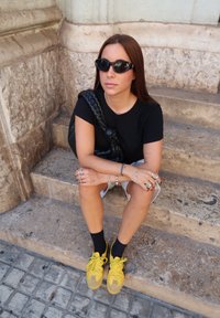 Camiseta preta, calções brancos, meias pretas e sapatos amarelos com detalhes em camurça. Uma bolsa trançada preta e óculos de sol completam o look.