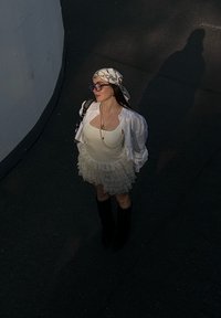 Vestido branco com folhos, corpete justo e saia em camadas. Botas negras até ao joelho, óculos de sol oversized e um lenço estampado complementam o visual.