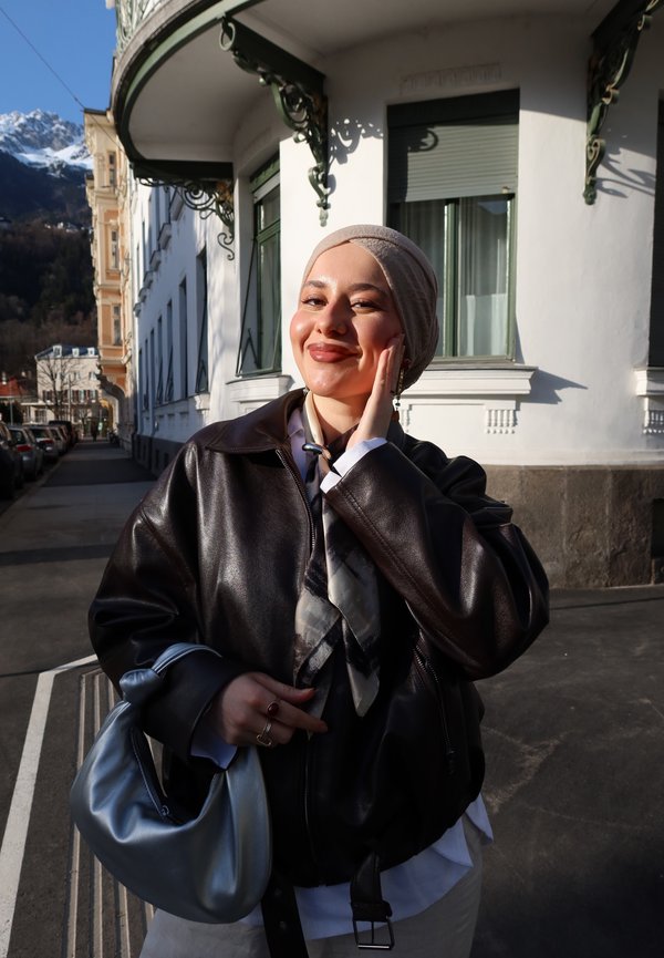 Młoda kobieta w beżowej chustce na głowie i ciemnej skórzanej kurtce uśmiecha się, trzymając srebrną torebkę na słonecznej ulicy z budynkami i górami w tle.