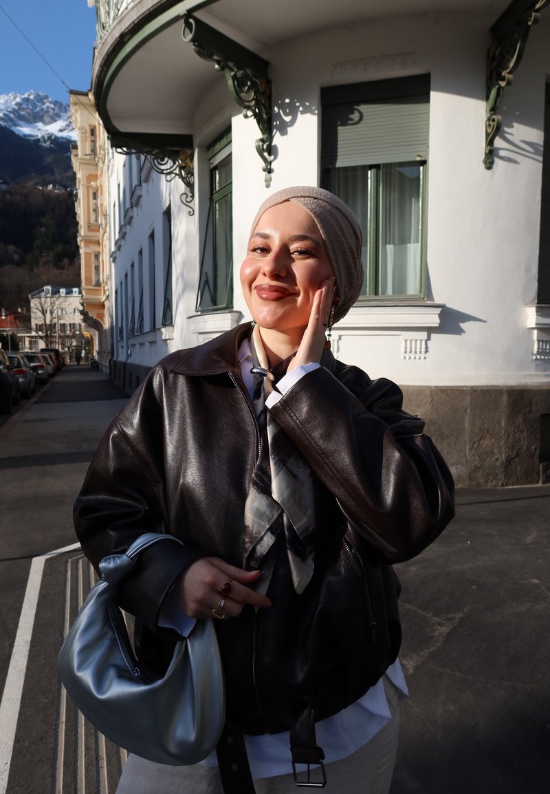 Junge Frau mit beigem Kopftuch und dunkler Lederjacke lächelt und hält eine silberne Handtasche auf einer sonnigen Straße mit Gebäuden und Bergen im Hintergrund.