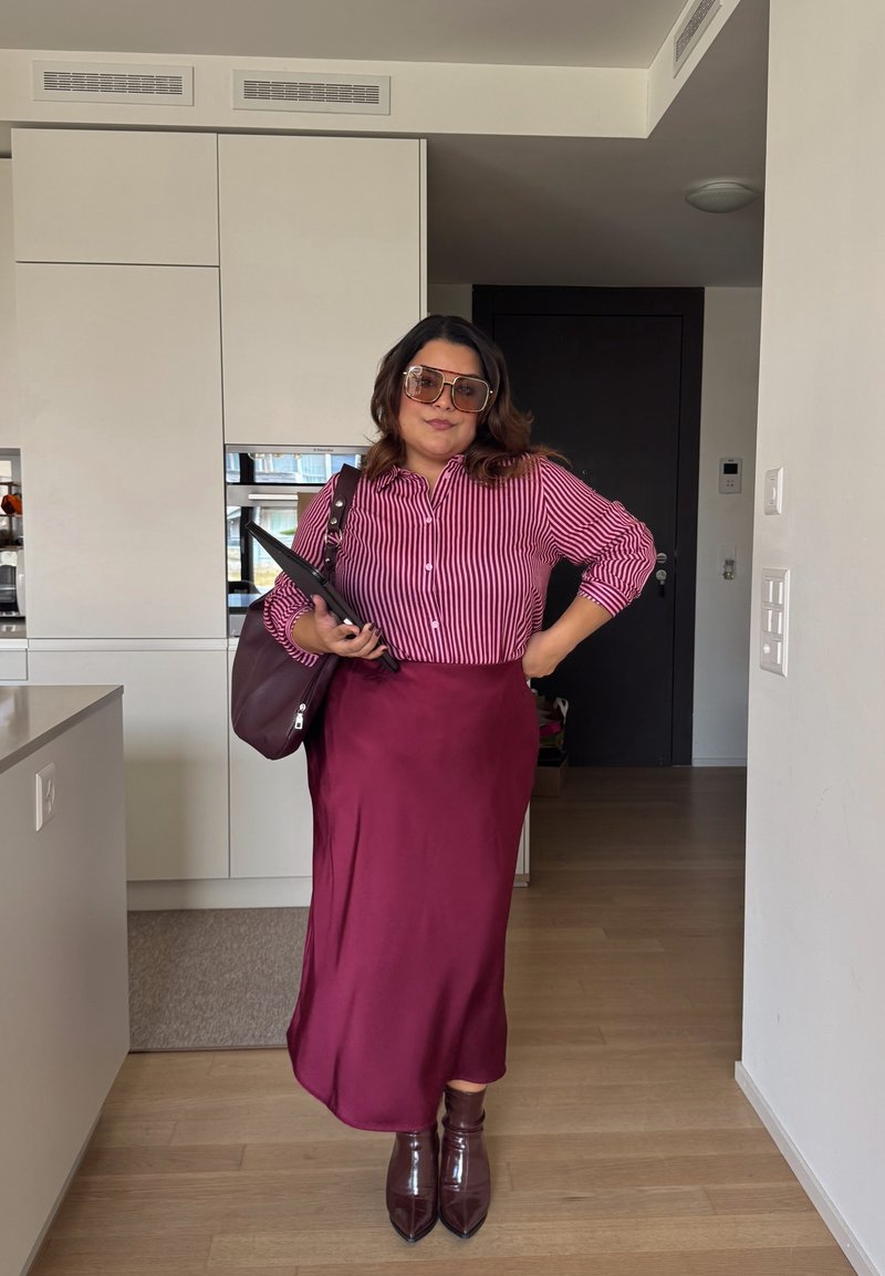 Jupe midi en satin bordeaux associée à une chemise boutonnée rayée rose et blanche et des bottes à talons marron foncé. Grandes lunettes de soleil et un sac à main.