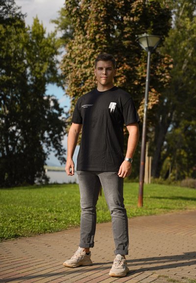 Camiseta negra con diseño gráfico blanco, jeans grises y zapatillas ligeras. El fondo presenta vegetación y un poste de luz.