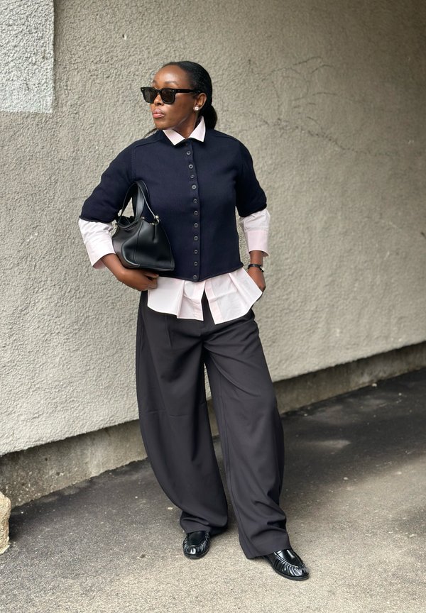 Donna con occhiali da sole neri, cardigan blu navy a maniche corte sopra una maglietta rosa, pantaloni neri larghi, che tiene una borsa nera, in piedi di fronte a una parete texturizzata.