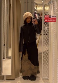 Μαύρο φουλάρι με φερμουάρ, μακριά σκούρα φούστα με ξεθωριασμένο σχέδιο και κρέμα φούτερ. Αντανακλαστικό φόντο με ράφια ρούχων.