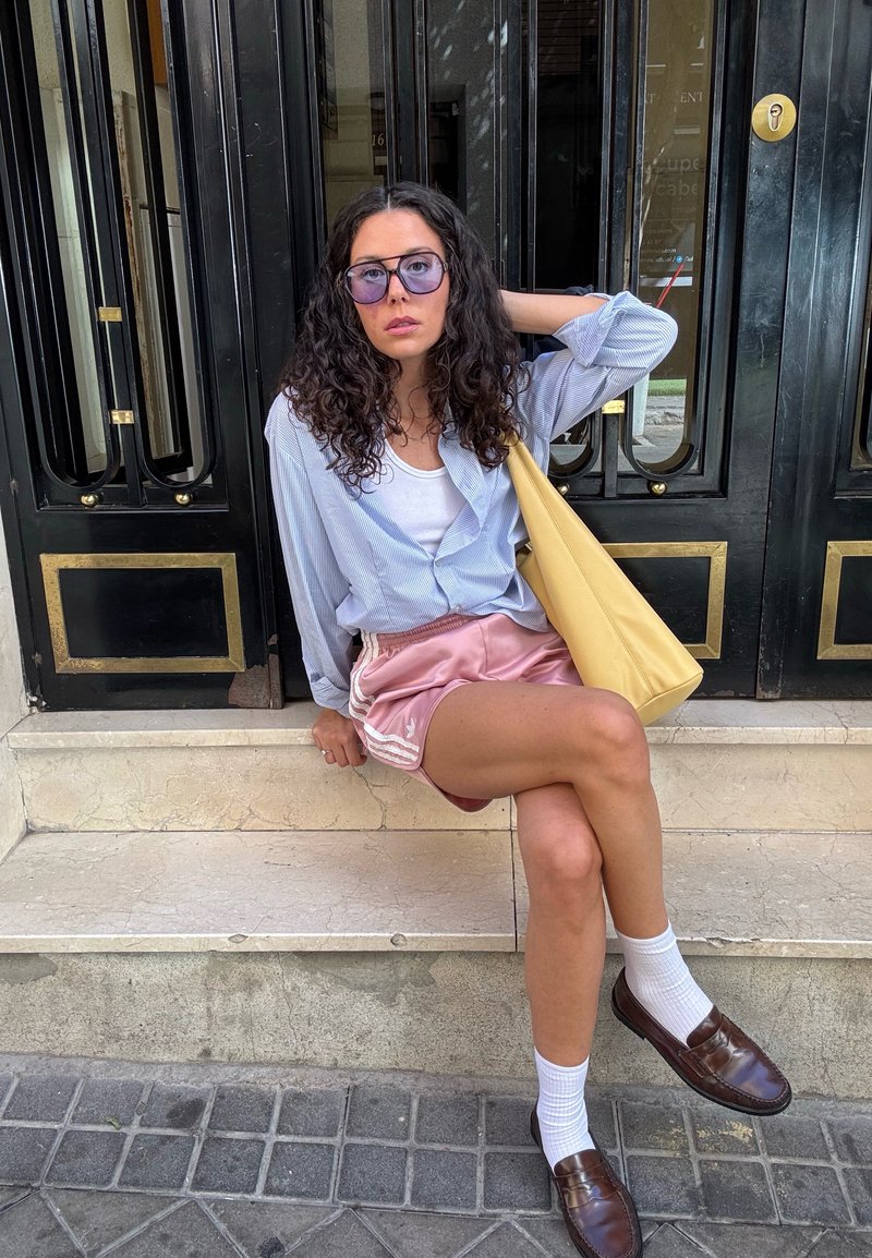Camicia a righe blu chiaro e bianca sopra un top bianco, pantaloni corti sportivi rosa, borsa gialla, calzini bianchi e mocassini marroni; seduto sui gradini.