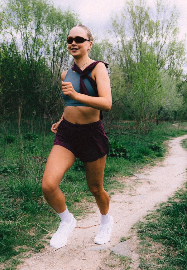 Vrouw jogt op een onverharde weg door een groene bosrijke omgeving, draagt een zonnebril, een blauw crop top, paarse shorts en witte sneakers.