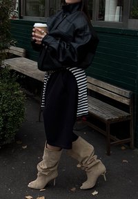 Casaco oversized em couro sintético preto, vestido preto com cinto riscado na cintura, botins altos beije e descaídos, segurando uma chávena de café.