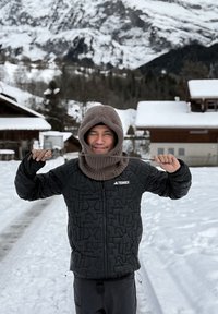 Veste matelassée noire avec fermeture éclair et logo, portée avec une capuche en tricot marron et une écharpe. Paysage enneigé avec des bâtiments en bois en arrière-plan.