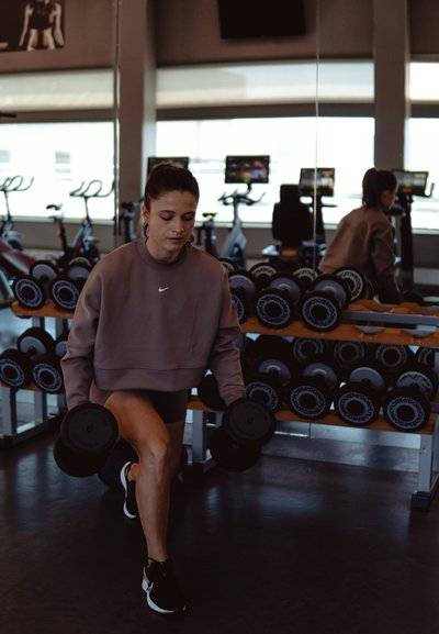 Mujer atlética con un sudadero gris de Nike y pantalones cortos negros está haciendo estocadas con dos mancuernas hexagonales en un gimnasio con equipo de fitness al fondo.