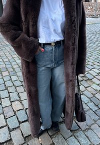 Personne portant un manteau en fausse fourrure brun foncé, un t-shirt blanc, un jean bleu, des chaussures noires, tenant un sac à main assorti, debout sur un pavé en pierre.