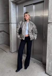 Metaalachtige zilveren bomberjack met een cropped wit topje, gecombineerd met zwarte wijde broek en zwarte hakken. Een kleine zwarte handtas is ook aanwezig.