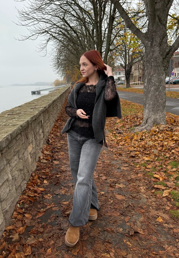 Grå genser over en svart, teksturert topp, kombinert med grå vide jeans og brune støvler. Stien rundt er dekket av høstløv.