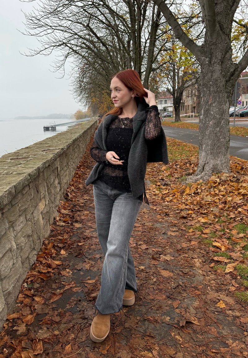 Pull gris par-dessus un haut texturé noir, associé à un jean gris évasé et des bottes couleur fauve. Le chemin environnant est couvert de feuilles d'automne.