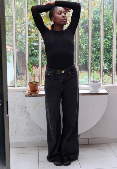 Mujer con suéter negro y pantalones estirando los brazos hacia arriba frente a una gran ventana con macetas en el alféizar.