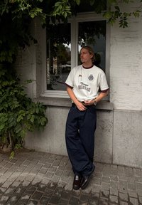 Muškarac sa svijetlom kosom leži uz bijelu ciglu pod zidom prekrivenim zelenim lozama, odjeven u bijelu majicu s natpisom "Berlin", tamne široke hlače i smeđe cipele.