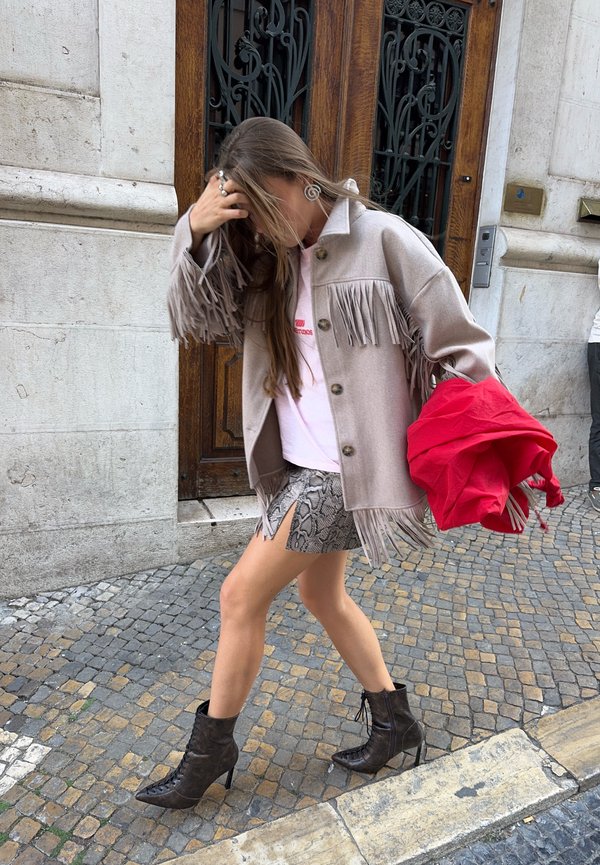 Mujer caminando por una calle empedrada con chaqueta de flecos, falda de piel de serpiente, botas negras de tacón, sosteniendo una chaqueta roja, cabello cubriendo el rostro.