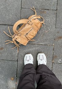 Läderhandväska i tan med flätad handtag, flera dragkedjefickor och dekorativ frans. Ligger på grå stengolv, nära sneakers.