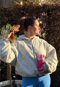 Jeune femme en sweat à capuche gris et leggings bleus tenant une bouteille d'eau rose Stanley, tirant sa queue de cheval en arrière, debout en extérieur près des plantes.