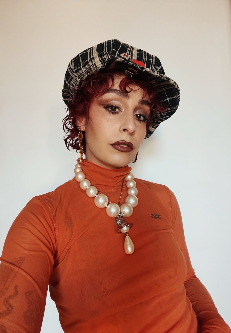 Personne aux cheveux rouges bouclés courts porte une casquette noire à carreaux, un col roulé orange, un grand collier de perles et une boucle d’oreille pendante, posant devant un fond uni.