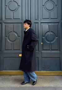 Jeune homme en long manteau noir, jean bleu et chaussures noires, debout de profil contre de grandes portes en bois foncé ornées de motifs circulaires.