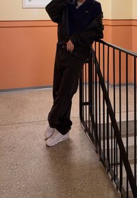 Musta nahkatakki tummansinisen poolopaidan päällä, väljät mustat housut ja valkoiset lenkkarit. Kohtauksessa on portaikko ja teksturoitu lattia.