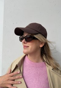 Brun caps, overdimensjonerte svarte solbriller, rosa strikkegenser og en beige trenchcoat. Myke teksturer og uformelle designelementer.