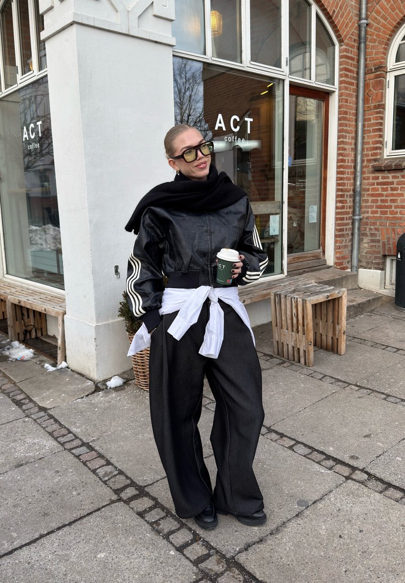Femme habillée d'une veste en cuir noire et d'un pantalon large tenant une tasse de café devant le café ACT, avec une façade en brique et en verre.