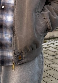 Harmaa pommikauppa takki, jossa on uurretut hihansuut, vetoketjullinen tasku ja ruudullinen paita alla. Pehmeä rakenne, yhdistettynä harmaisiin collegehousuihin.