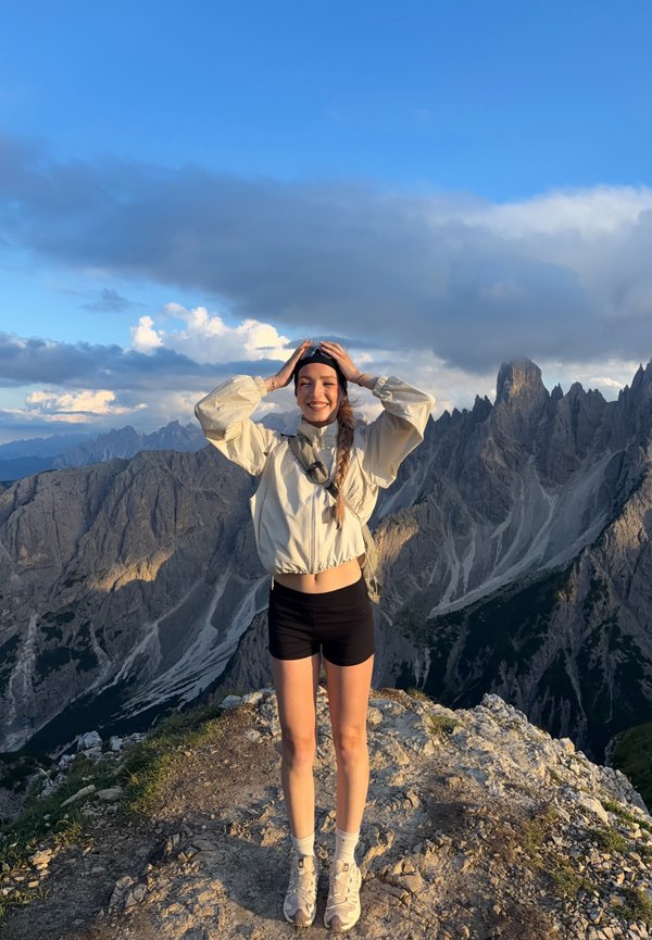 Jeune femme en short et veste debout au sommet d'une montagne rocheuse avec des falaises dentelées et un ciel nuageux en arrière-plan, souriante avec les mains sur la tête.