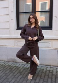 Jeune femme portant une tenue marron, des lunettes et des chaussures beige, posant avec une jambe levée devant un bâtiment clair avec une fenêtre.