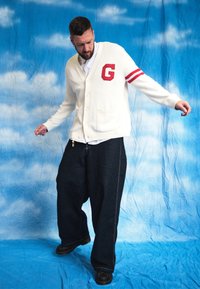 White cardigan featuring red stripes and a 'G' emblem, teamed with loose dark denim trousers. The model is wearing a white shirt underneath and is positioned against a blue backdrop.