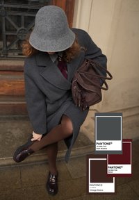 Personne portant un manteau gris et un chapeau, ajustant des chaussures marron foncé, tenant un sac marron avec des lunettes de soleil, debout sur des marches en pierre près d'une porte en bois.