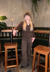 Brown striped blazer over a black top, teamed with brown trousers and black shoes. Standing next to wooden bar stools and green seating in an indoor setting.