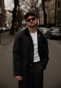 Manteau long noir avec un col texturé, t-shirt blanc et pantalon sombre. Lunettes de soleil noires et bijoux argentés complètent la tenue. Cadre urbain en extérieur.