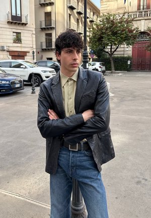 Joven con cabello rizado y oscuro, vistiendo una chaqueta de cuero negra y una camisa beige, de pie con los brazos cruzados en una calle urbana cerca de coches estacionados.