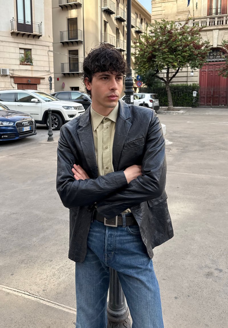 Young man with curly dark hair wearing a black leather jacket and beige shirt stands with arms crossed on urban street near parked cars.