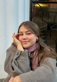 Cappotto di lana grigia con dettagli in bottoni, sciarpa rosa chiaro, capelli lunghi e scuri, seduta vicino a una colonna bianca, sfondo illuminato dolcemente.