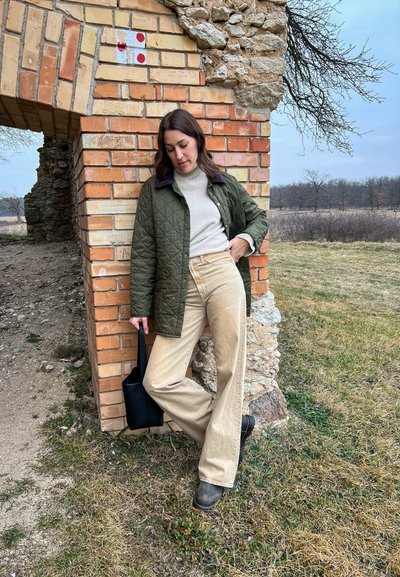 Chaqueta acolchada verde oliva, suéter de cuello alto crema, pantalones beige de patanla, bolso de mano negro y botas oscuras; de pie junto a una pared de ladrillo con acentos de piedra.