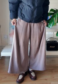 Henkilö, joka on pukeutunut mustaan untuvatakkiin, leveisiin beigeihin housuihin, joissa on valkoiset raidat, sekä ruskeisiin loafereihin ja valkoisiin sukkiin, seisoo sisätiloissa puulattialla.