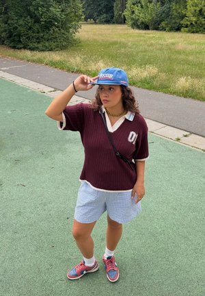 Top de punto oversized en color burdeos con detalles en blanco, pantalones cortos a rayas en azul y blanco, y gorra azul. Lleva zapatillas en rojo y azul con calcetas blancas.